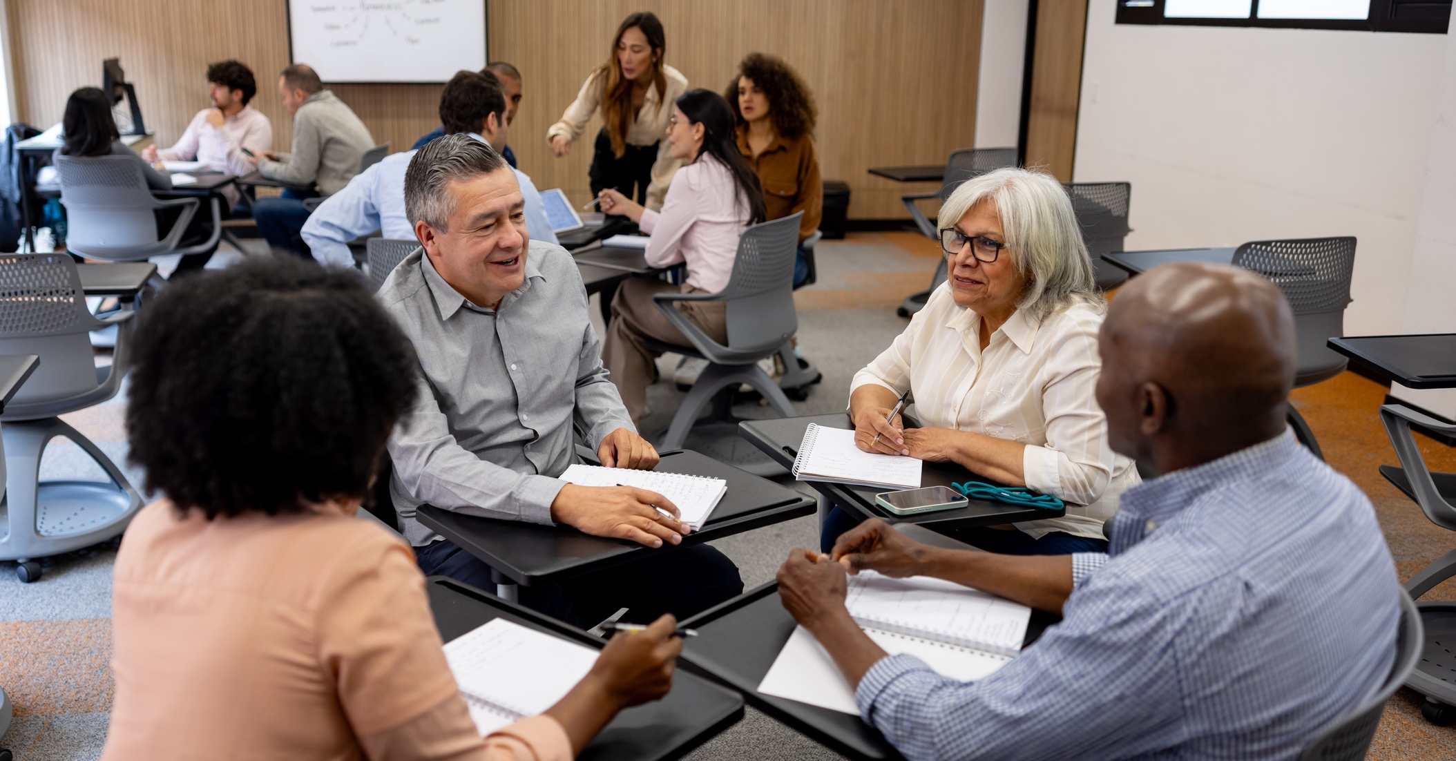 A diverse group of adults in a classroom setting sitting at desks, engaged in a discussion.