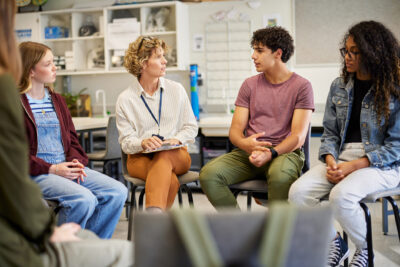 A teacher and four students sit in a circle in a classroom, engaged in a discussion.