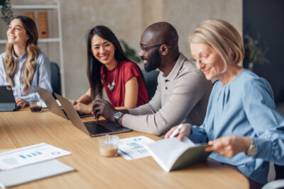 Four colleagues sitting at a wooden conference table with laptops and documents during a meeting.