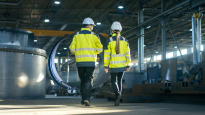 Two factory workers in hard hats and high-visibility jackets walking through an industrial manufacturing facility.
